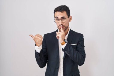 Beyaz arka planda dikilen yakışıklı İspanyol adam sessiz olmak istiyor. Parmakları dudaklarında, elleri yana doğru işaret ederek. sessizlik ve gizli konsept. 