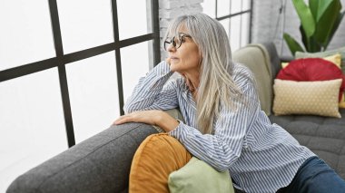 Düşünceli, olgun bir kadın. Gri saçlı. Rahat bir evin içindeki kanepede dinleniyor..