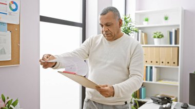 Süveter giyen orta yaşlı bir İspanyol, belgeleri parlak bir ofis ortamında inceleyerek profesyonelliği ve odaklanmayı resmediyor..