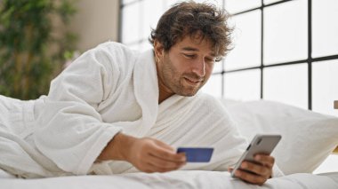 Bornoz giyen genç İspanyol adam yatak odasında akıllı telefon ve kredi kartıyla alışveriş yapıyor.
