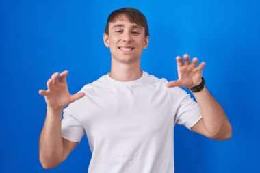 Mavi arka planda duran beyaz tenli sarışın adam komik bir şekilde gülüyor kedi gibi pençe hareketi yapıyor, saldırgan ve seksi bir ifade. 