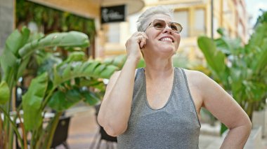Orta yaşlı, kır saçlı, gülümseyen, kendine güvenen bir kadın parkta akıllı telefondan konuşuyor.