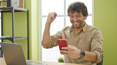 Genç İspanyol iş adamı ofiste akıllı telefon kullanarak kutlama yapıyor.