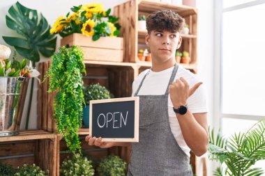 Çiçekçide çalışan İspanyol bir genç elinde açık tabela, başparmağıyla yan tarafı işaret ediyor, ağzı açık gülümsüyor. 