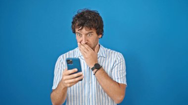 Akıllı telefon kullanan İspanyol genç adam mavi arka plana şaşırdı