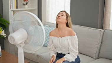 Kanepede oturan genç sarışın kadın evde vantilatör kullanarak sıcaktan acı çekiyor.