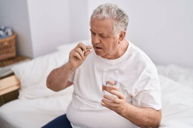 Orta yaşlı, kır saçlı, hap içen yatak odasında oturmuş su içen bir adam.