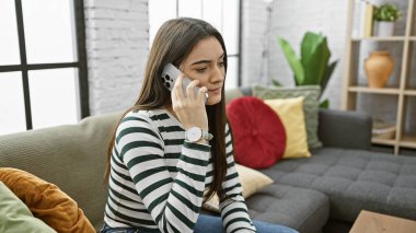 Kanepede oturan, çiçekli, rahat bir oturma odasında akıllı telefondan konuşan genç İspanyol kadın..