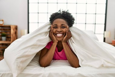 Afro-Amerikalı kadın, yatakta kendinden emin bir şekilde yatarken gülümsüyor.