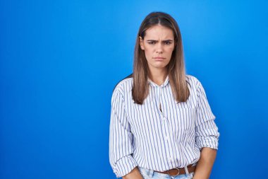 Mavi arka planda duran Latin kökenli genç bir kadın şüpheci ve gergin, sorun yüzünden surat asıyor. negatif kişi. 