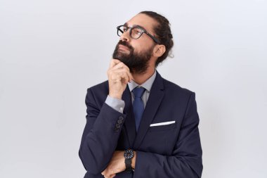 Hispanic man with beard wearing suit and tie with hand on chin thinking about question, pensive expression. smiling with thoughtful face. doubt concept. 