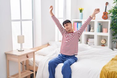 Young hispanic man waking up stretching arms at bedroom