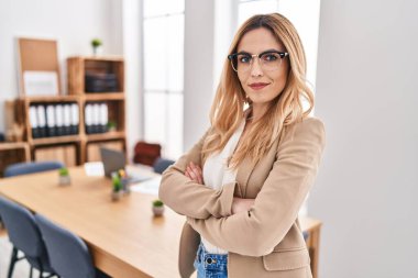 Genç, sarışın, iş kadını, gülümseyen, kendine güvenen, kollarını kavuşturup ofiste el kol kola duran.