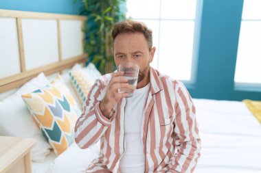 Middle age man drinking glass of water sitting on bed at bedroom