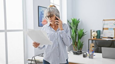 Gri saçlı, doküman okuyan ve modern bir ofiste telefonla konuşan olgun bir kadın..