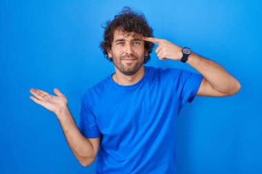 Mavi arka planda dikilen İspanyol genç adam avuç içinin açık olmasından rahatsız ve kafası karışmış bir şekilde fotokopi alanını gösteriyor ve alnını işaret ediyor. Bunu bir düşün.. 