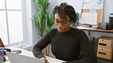 Rastalı saçlı, modern ofis ortamında dizüstü bilgisayarda çalışan odaklanmış Afrikalı Amerikalı kadın..