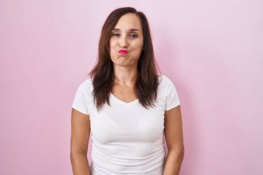 Orta yaşlı esmer kadın pembe arka planda durmuş komik suratlı yanakları şişiriyor. Havayla şişmiş ağız, çılgın bir ifade.. 