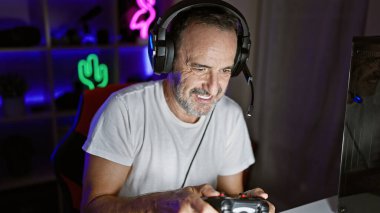 Gri saçlı orta yaşlı bir adamın duygusal akışı, oyun dünyasını bir joystick ile sallıyor, oyun odasında gecenin derinliklerine doğru gülümsüyor.