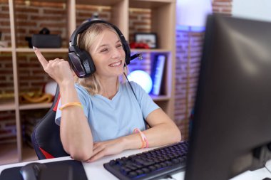 Video oyunları oynayan, kulaklık takan, yüzünde kocaman bir gülümseme olan beyaz bir kadın kameraya el ile işaret ediyor.. 