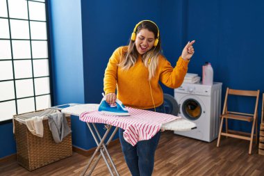 Young beautiful plus size woman listening to music ironing clothes at laundry room