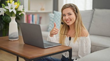 Laptop ve kulaklık kullanan genç sarışın kadın evde el hareketi yapıyor.