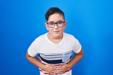 Mavi arka planda ayakta duran genç İspanyol çocuk midesi üstünde çünkü hazımsızlık, acı verici bir hastalık kendini iyi hissetmiyor. Ağrı kavramı. 