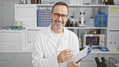 Gri saçlı mutlu orta yaşlı bir adam, kendine güvenen bir bilim adamı, elinde bir not panosuyla laboratuarında neşeyle çalışıyor.