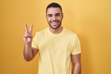 Sarı arka planda dikilen genç İspanyol adam iki numarayı gösterip işaret ederken kendinden emin ve mutlu bir şekilde gülümsüyor.. 
