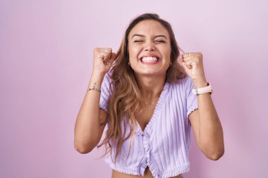 Pembe arka planda duran genç İspanyol kadın başarı için heyecanlı kolları havada ve gözleri kapalı zaferi kutlarken gülümsüyor. Kazanan konsept. 