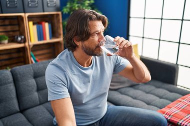 Orta yaşlı bir adam evdeki kanepede oturup su içiyor.