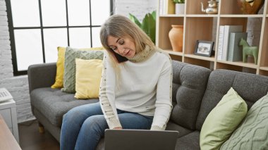 Konforlu bir oturma odasında birden fazla iş yapan bir kadın, dizüstü bilgisayarla çalışırken telefonla konuşuyor..