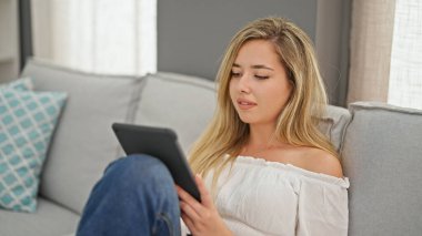 Young blonde woman using touchpad sitting on sofa at home