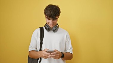 Havalı, genç İspanyol bir adam, mutlu öğrenci sırt çantalı, akıllı teknoloji yaşam tarzı yayıyor, telefonda mesajlar yazıyor. İzole edilmiş sarı bir arka planda kulaklıkla müzik dinlemekten hoşlanıyor..