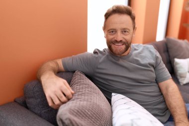 Orta yaşlı bir adam evdeki koltukta otururken kendinden emin gülümsüyor.