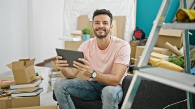 Yeni evindeki koltukta oturan dokunmatik ped kullanan İspanyol genç adam.