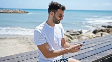 Young hispanic man using smartphone with serious expression at seaside