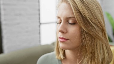 Kafkasyalı, kısa sarı saçlı yetişkin bir kadın, sakin bir oturma odasında bir kanepede oturuyor..