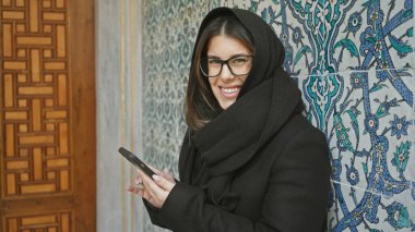 İstanbul 'da süslü bir Türk fayansına karşı akıllı telefon kullanan gözlüklü ve başörtülü bir kadın..