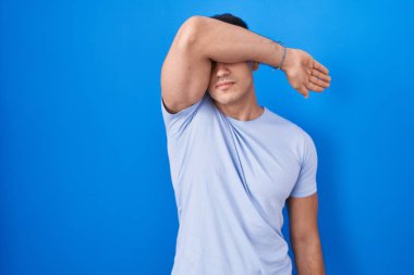 Mavi arka planda dikilen genç İspanyol adam gözlerini kollarıyla kapatıyor, ciddi ve üzgün görünüyor. Görme, saklama ve reddetme konsepti 