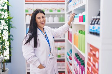Genç ve güzel İspanyol kadın eczacı gülümseyerek eczanede kendine güvenen ürünleri raflarda tutuyor.