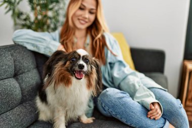 Beyaz bir kadın, evinde köpekle kanepede otururken kendinden emin bir şekilde gülümsüyor.