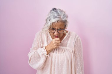Middle age woman with grey hair standing over pink background feeling unwell and coughing as symptom for cold or bronchitis. health care concept. 