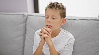 Sarışın çocuk evdeki kanepede dua ediyor.