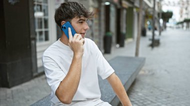 Gündüz Latin kökenli genç bir erkek, güneşli bir şehir sokağında ciddi bir telefon konuşmasına dalmış, havalı bir ifadeyle genç bir adamın hayat tarzı portresi, açık havada akıllı telefonuyla tutkuyla konuşuyor.