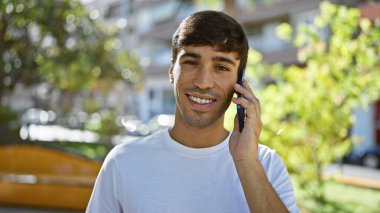 Güneşli parkta, kendine güvenen genç bir İspanyol adam duruyor, gülüşü ışıldıyor. Akıllı telefonundaki bir çağrıya kendini kaptırdı. Havalı bir resim. Açık havada saf mutluluğun portresi.