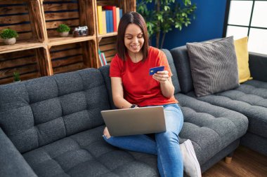 Evdeki kanepede oturan güzel İspanyol kadın dizüstü bilgisayar ve kredi kartı kullanıyor.