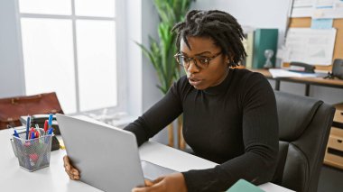 Rastalı saçlı, modern ofis ortamında dizüstü bilgisayarda çalışan odaklanmış Afrikalı Amerikalı kadın.