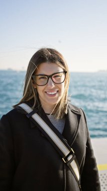 Güneşli bir günde deniz kenarında sıradan giysiler içinde gülümseyen ve gözlük takan genç bir kadın.