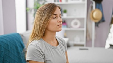 Çekici genç sarışın kadın, evde kanepede sakince otururken meditasyon yapıyor, endişeyi dengelemek için hava soluyor - evde endişeli bir ifade